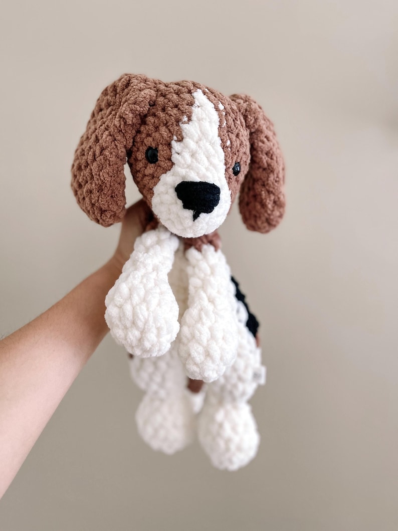 Pu&ograve; includere: Un giocattolo di peluche a uncinetto a forma di cane con pelliccia marrone, bianca e nera. Il cane &egrave; seduto con le zampe in aria.