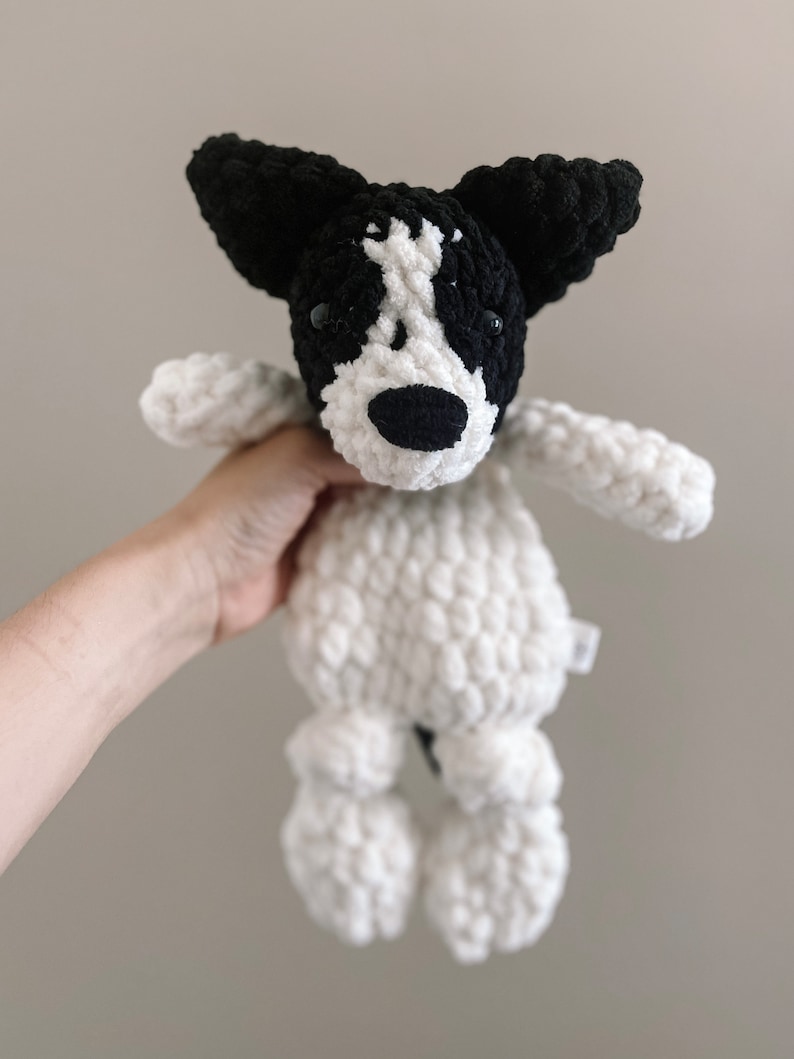 Pu&ograve; includere: Un giocattolo di peluche a forma di cane in crochet bianco e nero con occhi neri e naso nero. Il giocattolo ha una coda lunga e quattro zampe.