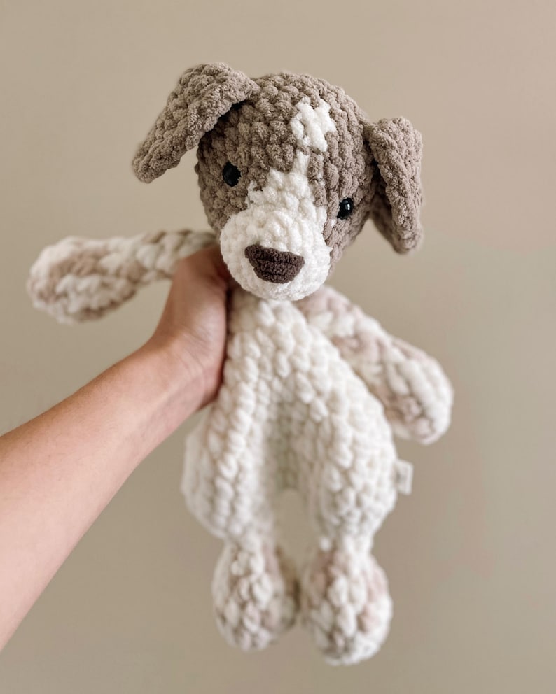 Pu&ograve; includere: Un giocattolo di peluche a forma di cane lavorato a uncinetto con filato marrone e bianco. Il cane ha una faccia bianca con occhi neri e un naso marrone.