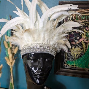 May include: A white feather headdress with a silver beaded band, displayed on a black mannequin head. The headdress features a crown of small, sparkling embellishments and a halo of long, elegant feathers. A silver necklace with dangling charms is also visible.