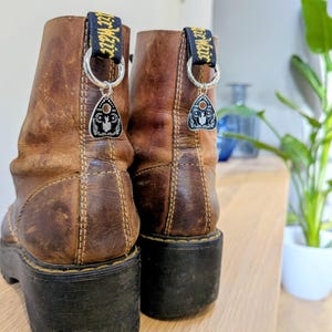 以下が含まれることがあります： 黒いソールと装飾的なチャームが付いた茶色の革ブーツ。チャームは黒とシルバーの縁取りで、プランシェットのデザインです。ブーツには黒と黄色のプルタブが付いています。