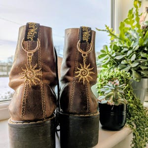 Könnte beinhalten: Paar braune Leder-Plateau-Stiefel mit goldener Sonnenstickerei und goldenen Ringakzenten. Die Stiefel haben schwarze Sohlen und ein abgenutztes Aussehen. Die Stiefel stehen auf einer weißen Fensterbank mit Pflanzen im Hintergrund.