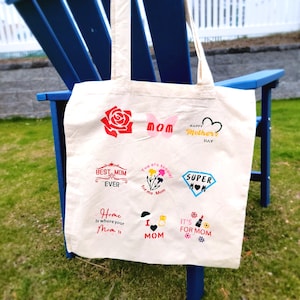 May include: A cream-coloured canvas tote bag featuring nine distinct graphic designs related to Mother's Day. The prints include a red rose, a pink butterfly with the word MOM, a heart, the text BEST MOM EVER, flowers, SUPER MOM, and various phrases like Home Is where your Mom Is and I LOVE MOM.
