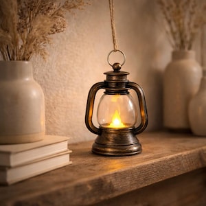 May include: A vintage-style lantern with a clear glass globe and a warm, flickering LED light. The lantern has a bronze finish and a metal handle for hanging. It is suspended by a natural fiber rope. The lantern is on a wooden shelf.