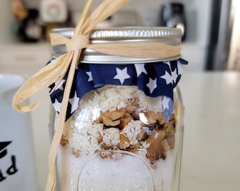 May include: A clear glass jar with a silver lid and a blue and white fabric tied around the top. The jar is filled with white sugar, chopped walnuts, and shredded coconut. A brown ribbon is tied around the jar.