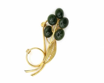 May include: A gold-tone brooch with a cluster of dark green glass stones in the shape of berries. The brooch is designed to look like a sprig of leaves.