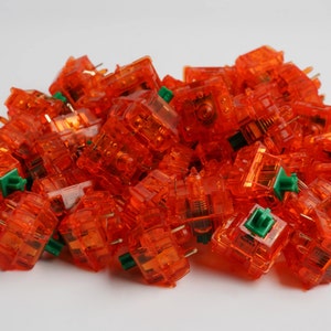 May include: A large pile of orange mechanical keyboard keycaps with green stems. The keycaps are translucent and show the internal mechanisms of the switches.