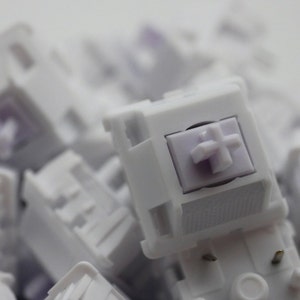 May include: A close-up of a white mechanical keyboard switch with a purple cross-shaped keycap. The switch is surrounded by other white switches.