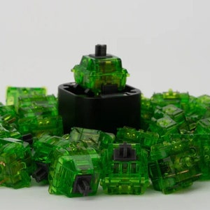 May include: A group of green mechanical keyboard keycaps, with one keycap in the centre that is slightly raised. The keycaps are made of transparent plastic and have a glossy finish.