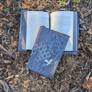 May include: A blue book cover with a textured, speckled finish featuring white embroidered dragon scales and two small dragon silhouettes. The cover is open, displaying the printed pages of a book with visible text on both sides of the open binding.