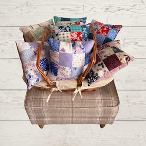 May include: A collection of square patchwork cushions featuring various floral and geometric patterns in shades of blue, purple, red, and white, arranged inside a woven wicker basket with a light-coloured fabric liner, resting on a plaid ottoman.