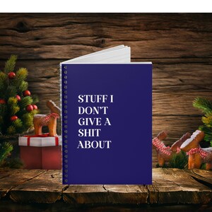 Puede incluir: Una libreta espiral azul con el texto "Stuff I Don't Give A Shit About" en letras blancas. La libreta está sobre una superficie de madera con un árbol de Navidad y adornos en el fondo.