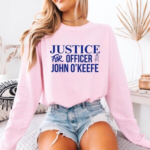 May include: A pink long-sleeve sweatshirt with the text "JUSTICE FOR OFFICER JOHN O'KEEFE" in blue lettering. The sweatshirt is worn by a person with long blonde hair and blue jeans.