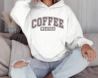 Puede incluir: Sudadera con capucha blanca con el texto "COFFEE WEATHER" en letras marrones.