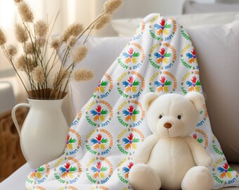 May include: A white plush teddy bear sits next to a white blanket. The blanket has a repeating pattern of colorful hands inside circles with the words "Teach, Understand, Accept, Love". A white vase with dried plants is in the background.