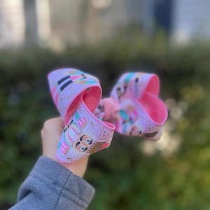 May include: A large, decorative hair bow featuring a pink and white pattern with a nutcracker design. The bow has a bright pink interior and is held up against a blurred green background.