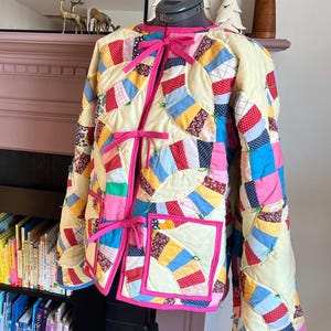May include: A vintage quilted jacket with a patchwork design in various colors, including pink, red, blue, yellow, and green. The jacket has a pink trim and ties at the front.