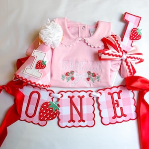 K&ouml;nnte beinhalten: Ein rosa Baby-Outfit-Set mit Erdbeermotiv. Das Set enth&auml;lt einen Strampler mit dem gestickten Namen "ISABELLA", eine Partyhut, eine Schleife, ein Banner mit der Aufschrift "ONE" und eine ausgeschnittene Zahl "1", alles mit rot-wei&szlig;em Vichy-Muster und Erdbeerakzenten.