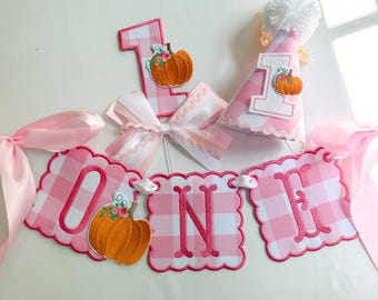 May include: A first birthday party set featuring a pink and white gingham banner spelling O-N-E with orange pumpkin appliqués, a number one cake topper with a pumpkin, and a matching party hat with a white pom-pom and a small pumpkin appliqué.