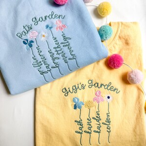 May include: Two embroidered sweatshirts, one light blue and one yellow, with floral designs and names. The light blue sweatshirt says "Pat's Garden" and the yellow one says "Gigi's Garden". Colorful pom-pom garland decorates the scene.