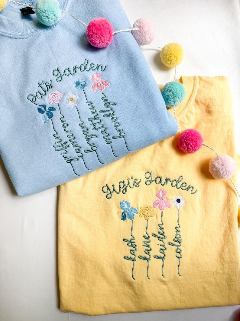 May include: Two embroidered sweatshirts, one light blue and one yellow, with floral designs and names. The light blue sweatshirt says "Pat's Garden" and the yellow one says "Gigi's Garden". Colorful pom-pom garland decorates the scene.
