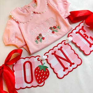 K&ouml;nnte beinhalten: Ein rosa Baby-Strampler mit wei&szlig;em R&uuml;schenkragen und floraler Stickerei. Der Name "MILLER" ist wei&szlig; gestickt. Ein Banner mit der Aufschrift "ONE" mit Erdbeere und rotem Vichy-Muster ist ebenfalls vorhanden.
