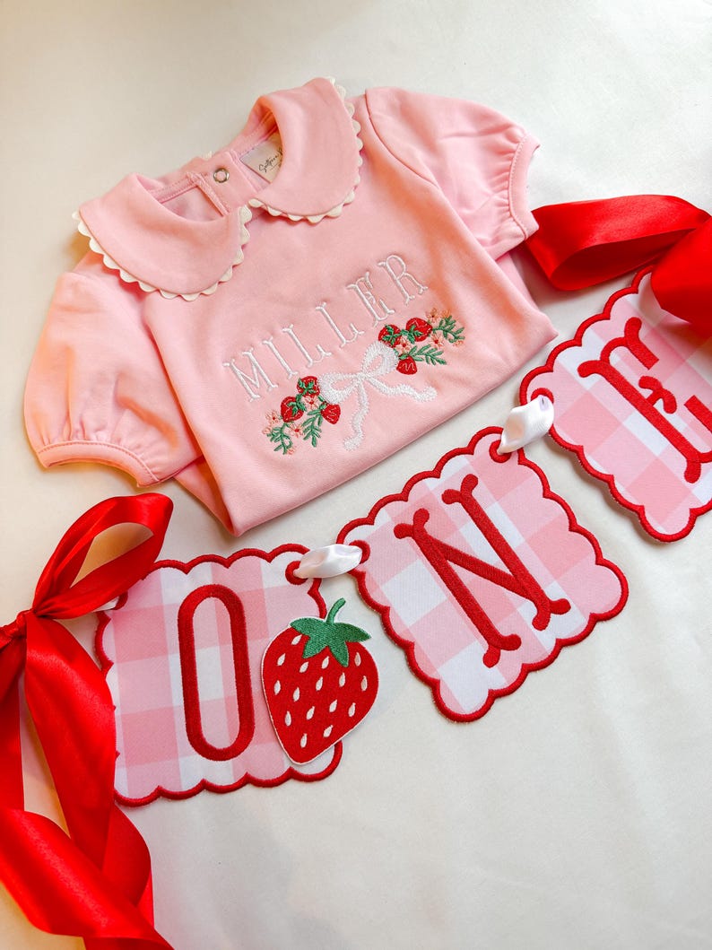 K&ouml;nnte beinhalten: Ein rosa Baby-Strampler mit wei&szlig;em R&uuml;schenkragen und floraler Stickerei. Der Name "MILLER" ist wei&szlig; gestickt. Ein Banner mit der Aufschrift "ONE" mit Erdbeere und rotem Vichy-Muster ist ebenfalls vorhanden.