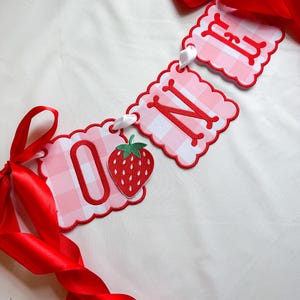 K&ouml;nnte beinhalten: Ein Banner mit dem Wort "ONE" in roten Buchstaben auf rosa Vichy-Karo-Anh&auml;ngern. Das Banner enth&auml;lt eine Erdbeer-Grafik und ist mit wei&szlig;em Band und roten Satin-Schleifen versehen.