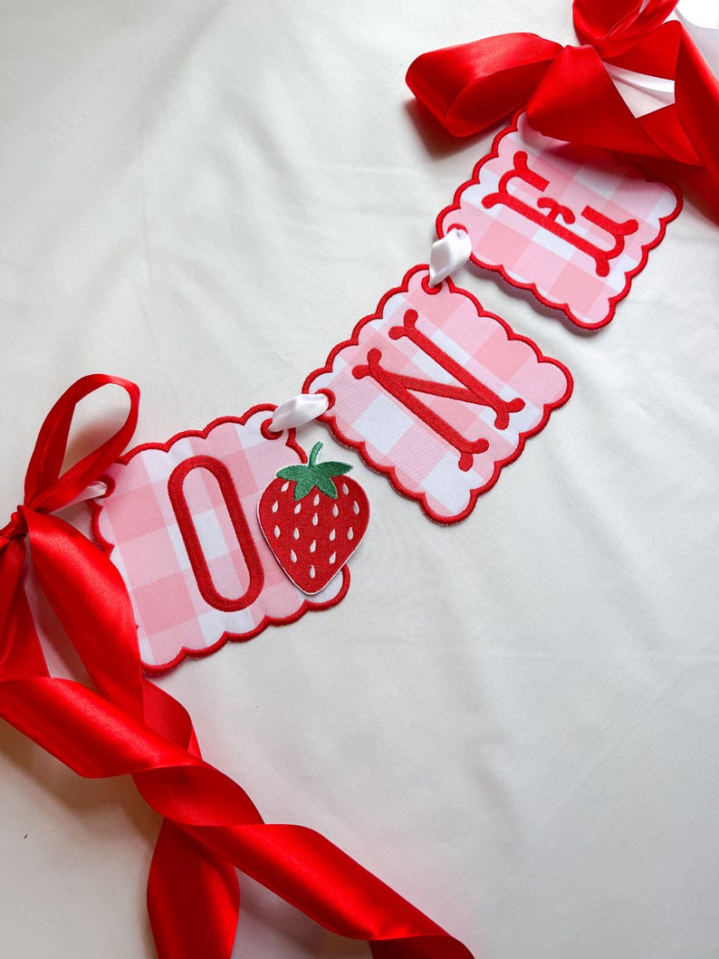 K&ouml;nnte beinhalten: Ein Banner mit dem Wort "ONE" in roten Buchstaben auf rosa Vichy-Karo-Anh&auml;ngern. Das Banner enth&auml;lt eine Erdbeer-Grafik und ist mit wei&szlig;em Band und roten Satin-Schleifen versehen.