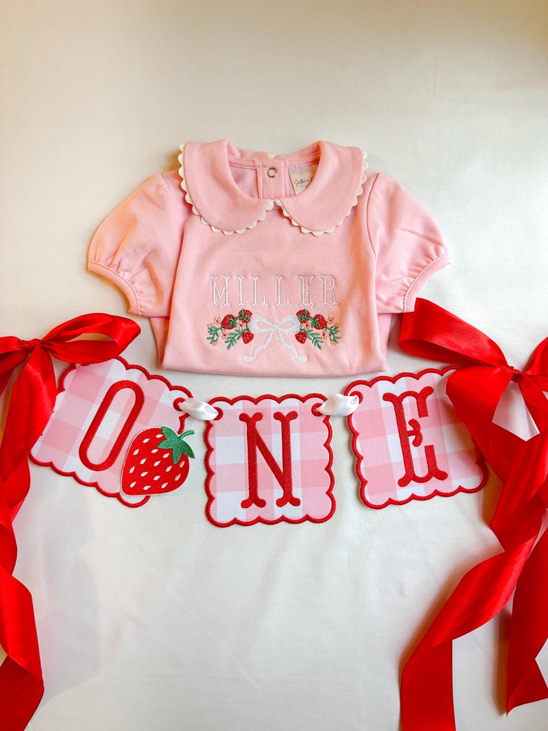 K&ouml;nnte beinhalten: Ein rosa Baby-Outfit mit einem R&uuml;schenkragen und dem Namen "MILLER" auf der Vorderseite gestickt. Ein Banner mit der Aufschrift "ONE" mit einer Erdbeere und einem rot-wei&szlig; karierten Muster ist ebenfalls vorhanden. Rote B&auml;nder sind auf jeder Seite gebunden.