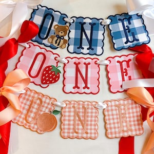 K&ouml;nnte beinhalten: Ein festliches Banner mit der Aufschrift "ONE" in drei verschiedenen Vichy-Mustern: Blau, Rosa und Pfirsich. Jeder Buchstabe befindet sich auf einer Karte mit gewelltem Rand. Die "O"-Karten zeigen einen Teddyb&auml;ren, eine Erdbeere und einen Pfirsich. Wei&szlig;e, rote und pfirsichfarbene B&auml;nder sind angebracht.