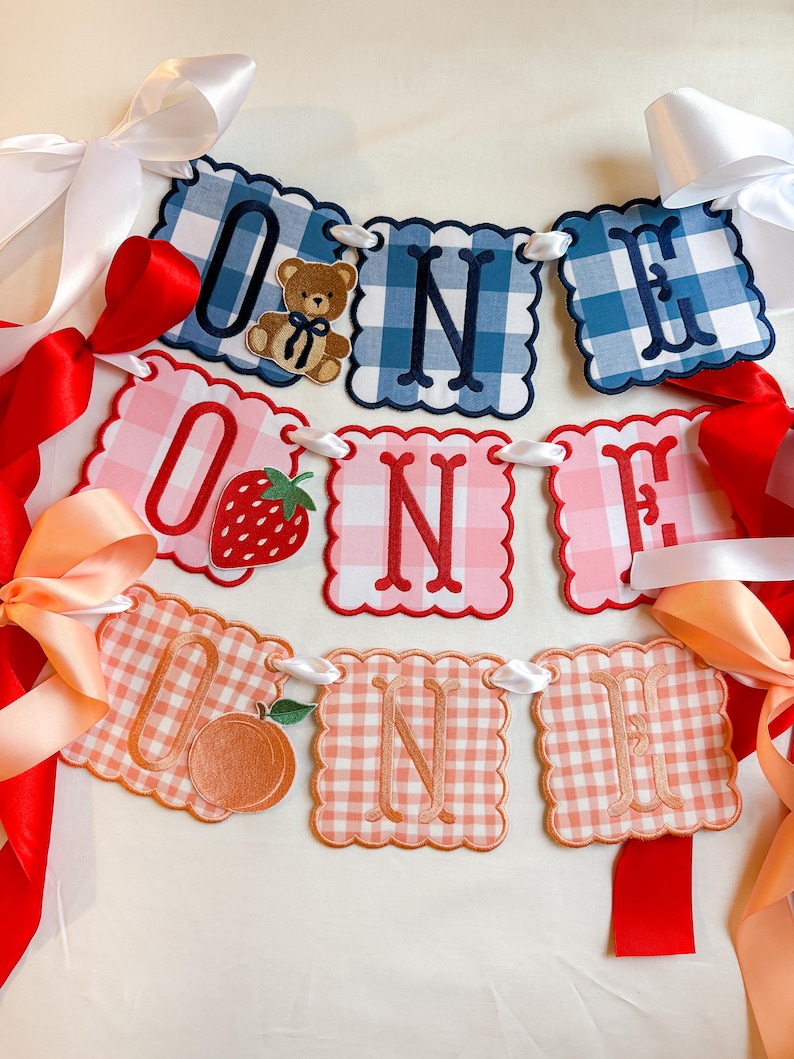 K&ouml;nnte beinhalten: Ein festliches Banner mit der Aufschrift "ONE" in drei verschiedenen Vichy-Mustern: Blau, Rosa und Pfirsich. Jeder Buchstabe befindet sich auf einer Karte mit gewelltem Rand. Die "O"-Karten zeigen einen Teddyb&auml;ren, eine Erdbeere und einen Pfirsich. Wei&szlig;e, rote und pfirsichfarbene B&auml;nder sind angebracht.