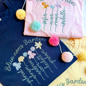 May include: Three embroidered t-shirts in navy blue, pink, and yellow. Each shirt features floral embroidery with names and the words "Mimi's Garden", "Blue-non's Garden", and "Gigi's Garden". A pom-pom garland is draped across the shirts.
