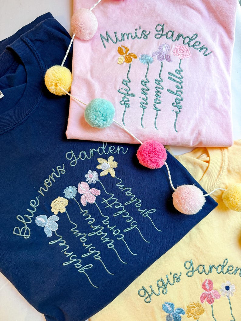 May include: Three embroidered t-shirts in navy blue, pink, and yellow. Each shirt features floral embroidery with names and the words "Mimi's Garden", "Blue-non's Garden", and "Gigi's Garden". A pom-pom garland is draped across the shirts.