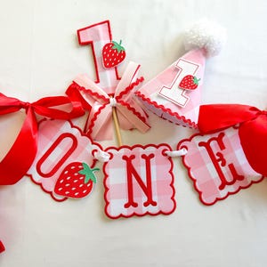 K&ouml;nnte beinhalten: Rosa-rote Geburtstagsdekorationen, darunter ein Banner mit der Aufschrift "ONE" und Erdbeeren. Enth&auml;lt die Zahl "1" und eine Partyh&uuml;tchen mit Bommel. Ideal f&uuml;r eine erste Geburtstagsfeier.