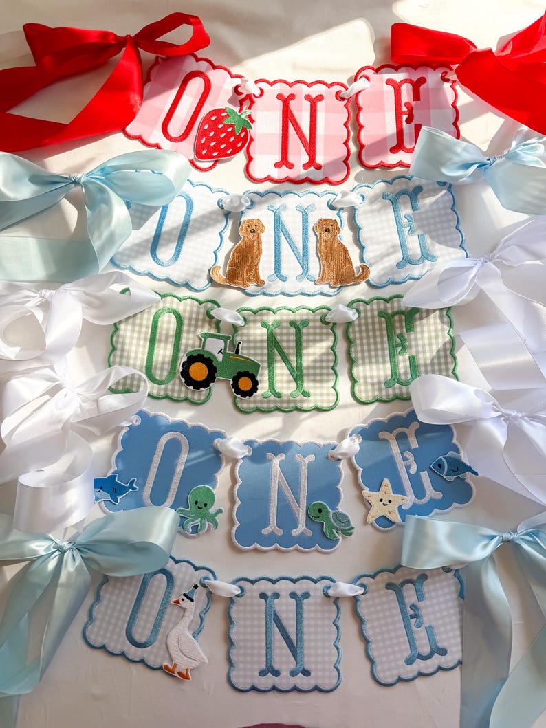 K&ouml;nnte beinhalten: Dekorative "ONE" Banner mit gewellten Kanten und verschiedenen Applikationen. Designs umfassen eine Erdbeere, Hunde, einen Traktor und Meerestiere. Die Banner sind mit wei&szlig;er Schnur verbunden und mit roten, wei&szlig;en und blauen B&auml;ndern akzentuiert. Geeignet f&uuml;r eine Feier.