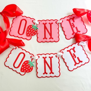 K&ouml;nnte beinhalten: Ein rosa-wei&szlig;es Banner mit dem Wort "ONE" in roten Buchstaben. Das Banner hat einen gewellten Rand und ist mit einer Erdbeergrafik verziert. Rote B&auml;nder sind angebracht.