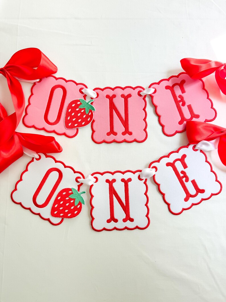 K&ouml;nnte beinhalten: Ein rosa-wei&szlig;es Banner mit dem Wort "ONE" in roten Buchstaben. Das Banner hat einen gewellten Rand und ist mit einer Erdbeergrafik verziert. Rote B&auml;nder sind angebracht.