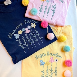 May include: Three t-shirts in navy blue, pink, and yellow, each embroidered with a floral design and the words "Blue-non's Garden", "Mimi's Garden", and "Gigi's Garden". Each shirt has a pom-pom garland.