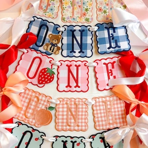 K&ouml;nnte beinhalten: Ein dekoratives Banner mit der Aufschrift "ONE" mit gewellten Kanten und verschiedenen Mustern, darunter Vichy, Blumenmuster und Obstdesigns. Das Banner ist mit wei&szlig;em Band aufgereiht und mit rosa, roten und pfirsichfarbenen Schleifen verziert.