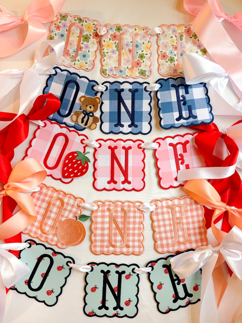 K&ouml;nnte beinhalten: Ein dekoratives Banner mit der Aufschrift "ONE" mit gewellten Kanten und verschiedenen Mustern, darunter Vichy, Blumenmuster und Obstdesigns. Das Banner ist mit wei&szlig;em Band aufgereiht und mit rosa, roten und pfirsichfarbenen Schleifen verziert.