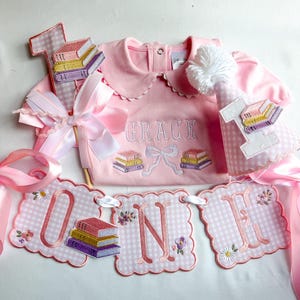 May include: A pink baby outfit set with a banner that says "ONE". The set includes a pink onesie with the name "GRACE" embroidered on it, a party hat, and a banner. The banner and hat have book and flower details.