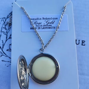 May include: A silver locket necklace with a round, white, solid perfume disc inside. The locket is open and the chain is visible. The card behind the necklace reads "Beaudin Botanique Rose Gold Solid perfume Made in Ladysmith"