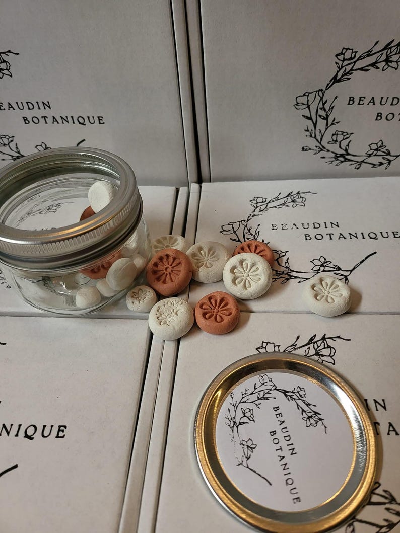 May include: A clear glass jar with a silver lid filled with small, white and brown round objects. Several round, floral-designed objects in white and terracotta colors are scattered around the jar. The packaging reads "BEAUDIN BOTANIQUE".