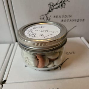 May include: A clear glass jar with a silver lid filled with white and peach-colored treats. The jar has a white label with the text "BEAUDIN BOTANIQUE" and a floral design. The jar is sitting on a white surface with the same logo.