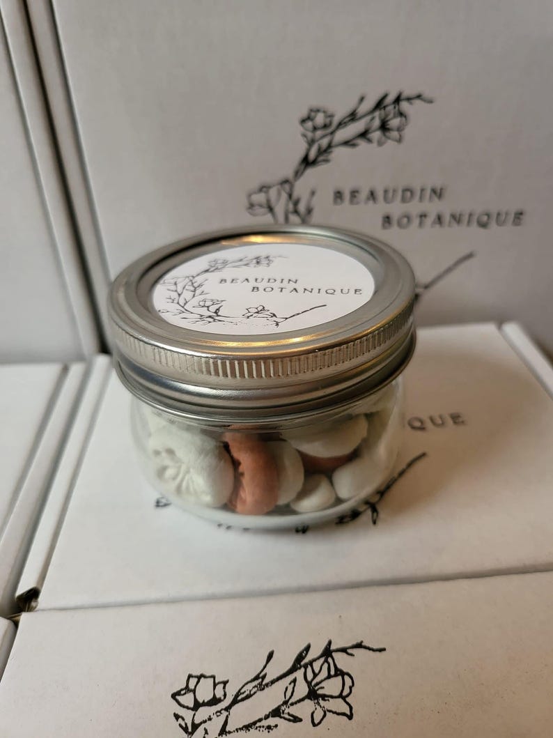 May include: A clear glass jar with a silver lid filled with white and peach-colored treats. The jar has a white label with the text "BEAUDIN BOTANIQUE" and a floral design. The jar is sitting on a white surface with the same logo.