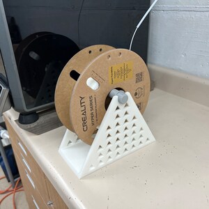 May include: A white 3D-printed filament spool holder with a triangular design, holding two brown filament spools. The spools are labeled "CREALITY HYPER SERIES". The holder is on a light-colored surface.