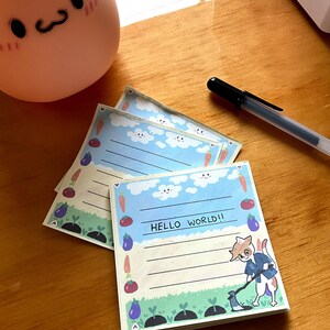May include: A stack of colorful notepads with a blue sky, white clouds, and a cartoon cat with a hoe in a garden setting. The notepad has lines for writing and the text "HELLO WORLD!!" is printed on the notepad.