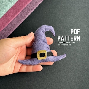 May include: A small, purple felt witch hat with a black band and gold buckle. The hat is held in a hand, with the text "PDF PATTERN + PHOTO AND TEXT INSTRUCTIONS" in the background. Felt fabric in shades of purple and green are in the upper left corner.