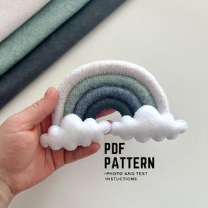 May include: A felt rainbow craft project with a white, green, grey, and dark grey colour scheme. The rainbow has two white felt clouds attached to the bottom. The text "PDF PATTERN +PHOTO AND TEXT INSTRUCTIONS" is printed below the rainbow.